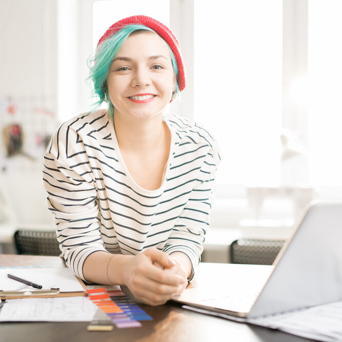 Portrait of contemporary creative young woman with mint-colored hair smiling happily looking and camera while standing at work table in sunlit design studio
